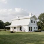 A stunning white barndominium featuring large windows and rustic charm amidst a lush landscape.